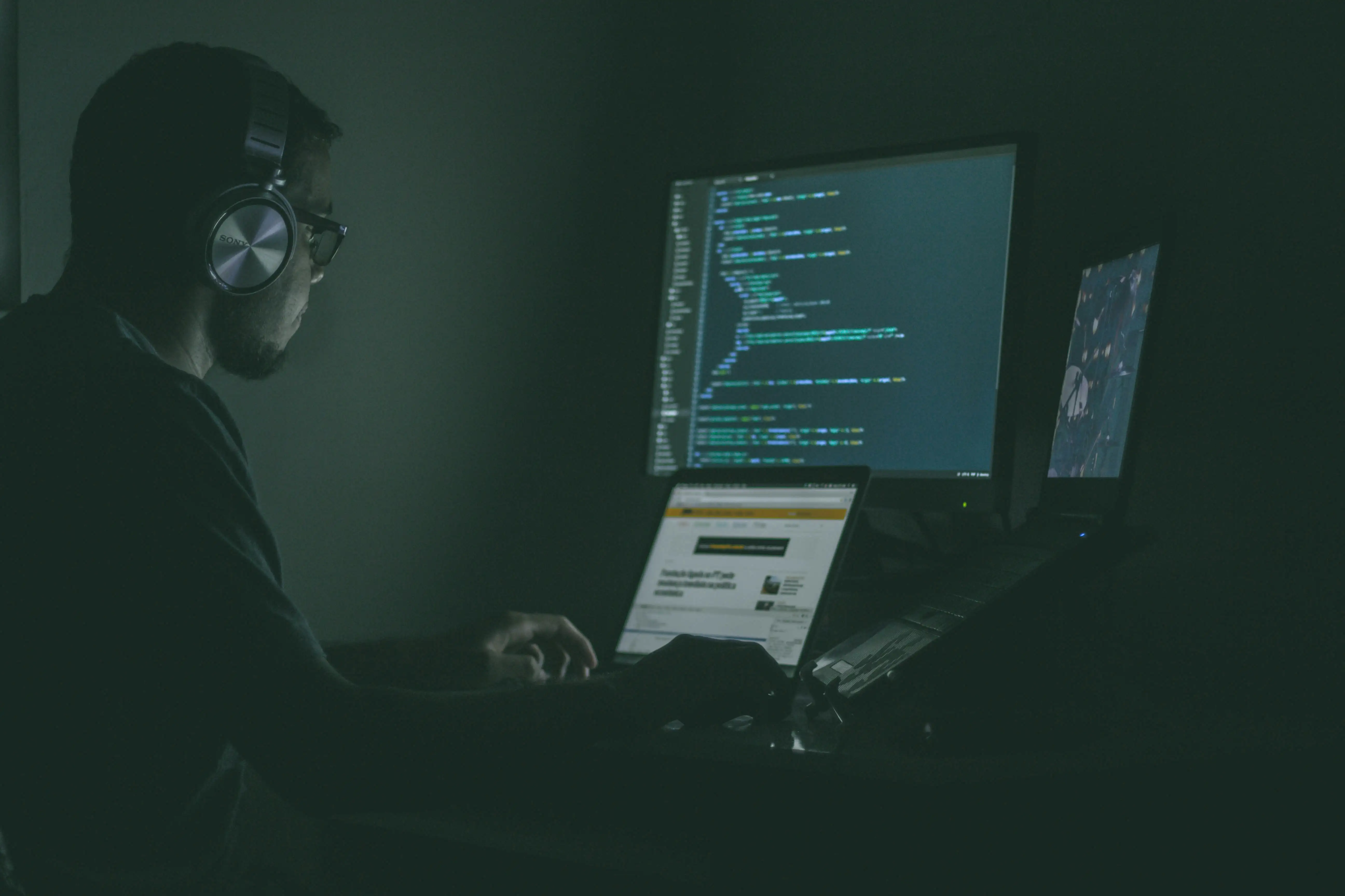 A man sitting on a desk with three displays wearing a headphone.